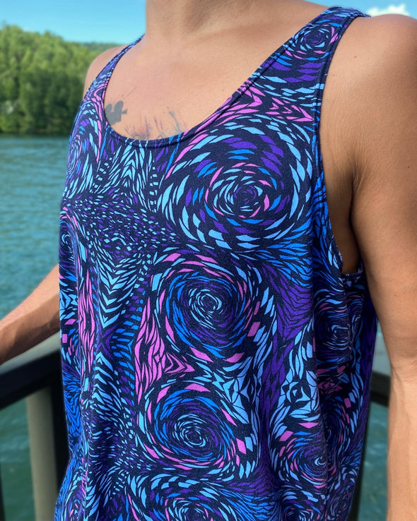 Mid shot photograph of a male model from the waist up in the foreground wearing a Bleace psychedelic tank top with pink, turquoise and purple swirls on it with Lake Rabun, Georgia in the background.